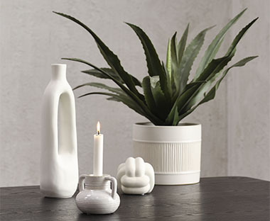 Bele dekoracije na stolu - vaza, saksija sa zelenom veštačkom biljkom, svećnjak i ukras