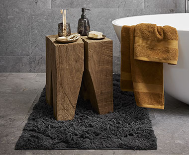 Siva prostirka za kupatilo na sivom podu i na njemu drveno postolje sa dodacima za kupatilo