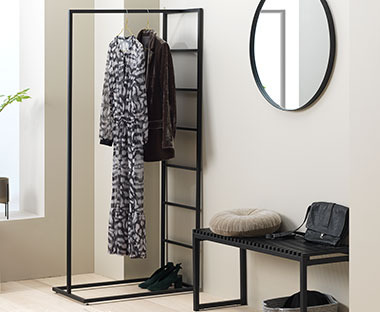 Crni metalni stalak za odeću u predsoblju pored crne klupe i crnog okruglog ogledala