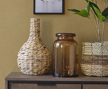 Vaza od pruća i braon staklena vaza pored korpe od pruća sa veštačkom biljkom