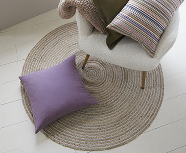 Okrugli juteni tepih bež boje na kome su fotelja i ljubičasti ukrasni jastuk