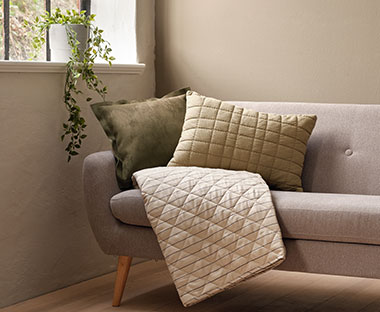 Sivi kauč sa jastucima i ćebetom neutralnih boja pored prozora