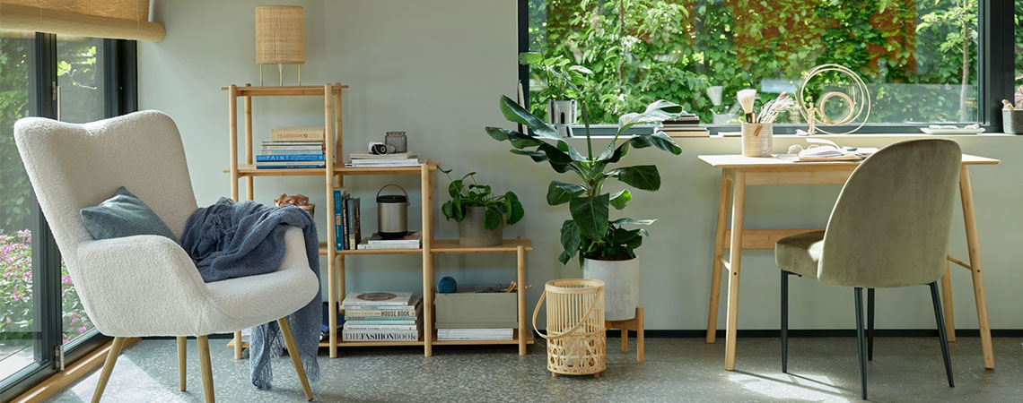 Siva fotelja kraj prozora i komoda od bambusa u pozadini. Bambusov fenjer na podu pored stola od bambusa i maslinasto-zelena stolica